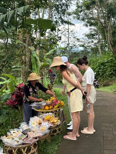 The image depicts a scene in Penebel, Indonesia, near the Jatiluwih rice terraces. Two fair-skinned women wearing summer attire—one in a yellow sundress and the other in a white top and beige shorts—stand on a concrete path and appear to be interacting with a local merchant. The merchant, wearing a straw hat and a floral-print shirt, sits surrounded by baskets filled with various snacks and souvenirs. The path is lined with lush vegetation, including banana trees and tropical plants. A parked scooter is visible in the background.