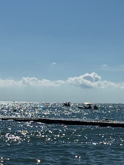 In Adlerskiy, Russia, on a bright, sunny day, the turquoise sea is calm and shimmering with reflections of sunlight. A dark, rectangular structure, possibly a pier or breakwater, runs horizontally across the middle ground of the image, separating the foreground and background waters. Several small boats, carrying passengers, are visible in the distance on the water. The sky above is a brilliant blue, dotted with fluffy white clouds.
