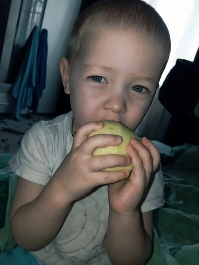 A young child with fair skin and blue eyes is holding and biting into a yellow fruit, possibly an apple or pear. The child is wearing a light-colored t-shirt. The background includes dark, undefined objects and a surface with blue and green cloth. The photograph appears to be taken indoors in an unknown location. The location of Yurgamysh, Russia is not ascertainable from visual inspection.