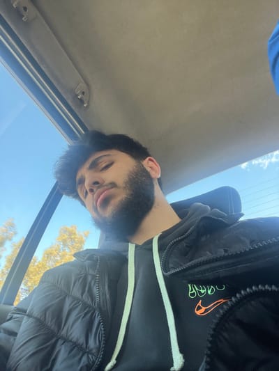 The image depicts a young adult male seated inside a vehicle. The male has dark, curly hair, a full beard, and is looking towards the camera with a slightly pouting expression. He is dressed in a black puffer jacket layered over a black hooded sweatshirt with light green drawstrings. An orange Nike swoosh logo and an additional green graphic are partially visible on the front of the sweatshirt.

The interior of the vehicle is visible, including a light gray headliner with two small plastic fixtures in the upper left. Through a window to the left of the subject, a clear blue sky with minimal white clouds is visible, along with the upper sections of trees displaying yellowish-brown foliage. The scene is brightly illuminated by natural daylight. No specific landmarks or unique architectural features are present in the visible background to definitively pinpoint the exact location within the city of Ashrafia, Syria.
