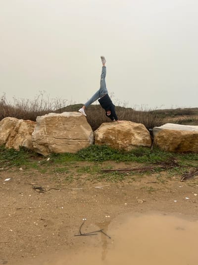 The image depicts a single individual performing an inverted pose on large boulders in Jisr az-Zarqa, Israel. The person, wearing a dark hooded jacket, light blue jeans, and white sneakers, has both hands on a central light-brown boulder. Their left leg is extended vertically upwards, while the right leg is bent at the knee, with the foot resting on an adjacent boulder to the right. The individual's face is visible, directed towards the camera, with a slight smile. Behind the rocks, dry, brownish scrub vegetation is present, transitioning into sparse green growth on the ground immediately behind the rocks. The foreground consists of muddy-brown earth with various pieces of debris, including dark tangled wires, white plastic, and dry plant matter. A large, irregularly shaped puddle of muddy water occupies the lower right corner of the foreground. The sky is uniformly overcast, presenting a light brownish-grey hue with no discernible features. The environment appears to be an undeveloped or natural area.