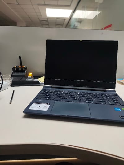 The scene depicts an unoccupied desk within an office environment in the city of Kangli, Uzbekistan. A dark-colored HP Victus gaming laptop is open on a light-colored desk, its screen remaining dark. The laptop features a non-English keyboard layout and visible stickers on the palm rest, including "Victus by HP Gaming Laptop" and an Intel Core i5 badge. To the left of the laptop, a black mechanical pencil rests horizontally on the desk. Further left, a black desk organizer unit holds various stationery items, next to which sits a small, amber-colored translucent object and a stack of yellow sticky notes. In the background, a light-colored office partition wall is visible, topped by a grid-patterned ceiling with illuminated rectangular fluorescent light fixtures. A glass partition on the right reflects these lights and shows a faint red shape beyond it. A partial view of a light-colored object, possibly a chair back, is visible in the extreme bottom foreground. No individuals are present.