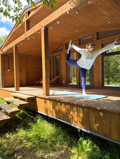 An energetic outdoor scene captures a young woman performing a dynamic pose on a wooden deck next to a modern cabin. The woman, with blonde hair and wearing a white long-sleeved top, dark blue leggings, and sunglasses, extends one leg high behind her while her arms are outstretched, resembling a split or an acrobatic dance move. She is positioned on a light blue yoga mat on the wooden deck.

The cabin behind her features extensive wooden siding and large, dark-framed windows that reflect the surrounding lush green trees and blue sky. String lights are strung along the wooden beams of the cabin's overhang, suggesting an evening ambiance feature. Below the deck, wooden stairs descend into an area abundant with vibrant green grass and foliage, indicating a natural, perhaps wild, environment.

The image is captured during bright daylight, evidenced by the strong shadows cast across the wooden surfaces, suggesting either morning or late afternoon sun. The weather appears clear and sunny. The overall setting combines modern architecture with a serene natural landscape, creating a peaceful yet active atmosphere. No visible text is present in the image.