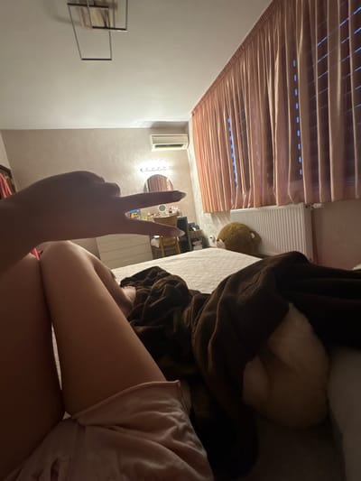 The image is a close-up shot of a person lying down, possibly in a bedroom. The person is making a peace sign with their hand, with their nails painted. They are wearing light-colored clothing. In the background, there is a window with blinds, a radiator, and what appears to be a stuffed animal. The lighting suggests it might be evening or nighttime. The overall scene is casual and intimate, hinting at a moment of relaxation or self-expression. There is no visible text in the image.