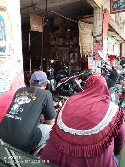 The scene takes place in a motorcycle repair shop in Kecamatan Haurgeulis, Indonesia. The shop is located in a densely populated area, with several buildings and a busy street. There are two people in the shop: a man wearing a black t-shirt with a monkey on it and a woman wearing a maroon headscarf with a white lace trim. The man is sitting on a stool, looking at a motorcycle. The woman is sitting on the floor, looking at the man. The shop appears to be a busy one, with several motorcycles parked around the shop and tools scattered about. The shop is messy, with papers and other debris scattered around. The shop is dimly lit, and the walls are painted in a dull, gray color. There are two motorcycles partially visible, one with a white and red color scheme and one with a black and red color scheme. The air inside the shop is thick with the scent of oil and gasoline.