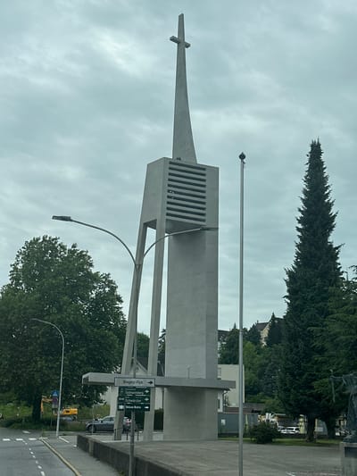 This is a photograph of a modern church tower with a prominent cross at the top, located in the city of Bregenz, Austria. The tower is made of light-colored concrete and features horizontal slats on its upper section. A street sign with multiple destinations is visible at the base of the tower, including "Bregenz-Fluh", "Gebhardsberg", "Schwarzer", and "Deluxe Bistro". A street lamp curves over the road in the foreground. Several trees, including a tall, dark evergreen tree, are present in the background. A grey sedan is parked on the right side of the road, and a yellow excavator is visible in the distance on the left. The sky is overcast.