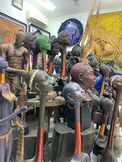 The image displays an indoor art gallery or studio located in Ikate, Nigeria, densely filled with a collection of sculptures and artworks. The environment features white walls, a white tiled floor, and overhead fluorescent lighting, with a wall-mounted air conditioning unit visible on the upper left.
The primary subjects are numerous sculptures, predominantly busts and full figures of humans, along with some animal representations. Multiple busts of African women with elongated necks are prominent in the foreground and mid-ground. These busts exhibit various dark skin tones, with one notably green-colored. Many feature copper-colored hoop earrings and some have copper bands or textured patterns on their elongated necks. One female bust in the mid-ground displays a yellow and white patterned headband.
Among the full figures, a muscular, dark brown male sculpture wearing a textured loincloth is visible on the left, holding a long, narrow object resembling a fish or spear. Another purple-grey female sculpture in the foreground wears a gold-colored top and skirt, holding a pole with a fish attached.
An animal head sculpture with large, spiraled, light brown horns, indicative of an antelope, stands behind the main group of busts. The sculptures are displayed on dark wooden pedestals and a circular, intricately carved wooden table in the mid-ground.
The background includes several wall-mounted artworks: a rectangular piece with textured brown and orange abstract forms on the far left, a circular blue artwork depicting the African continent outline with a profile head surrounded by textured elements, and a large, textured yellow/gold relief artwork with organic, possibly floral or vegetative, patterns on the right. No live individuals are present; all depicted entities are static artworks.