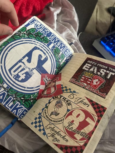 The image shows an open notebook with several stickers adhered to its pages. The notebook is lying on a surface that appears to be a bed or sofa.
The left page of the notebook features a large circular sticker with a blue and white emblem. The emblem contains the numbers "04" in large blue font, with a white "S" superimposed on the "0". The outer edge of the circle has green leaf-like designs. Above the emblem, the text "FÜR IMMER" is printed in blue uppercase letters. Below the emblem, the word "SCHALKE" is visible, also in blue uppercase letters. A small sticker of a Viking-like character with a red and white striped background is partially obscuring the main emblem.
The right page of the notebook contains several more stickers. One prominent sticker displays the text "BOYS FROM THE EAST" in large white letters against a dark red background. Below this text are two crests, one with red and white stripes and a crown, and another with a white horse on a red background. The text "FC. TWENTE" and "Twente Enschede" are also visible on this sticker.
Below the "BOYS FROM THE EAST" sticker, a circular sticker with the number "87" in a red circle with white outlining is visible. The text "Konnuti" is written in white script within the red circle. To the left of the "87" sticker, another circular sticker features a cartoon character with a baseball cap and the text "ULTRAS GELSENKIRCHEN". The phrase "15 Years" is written in script above this sticker.
The notebook pages are lined, and the overall impression is that of a collection of football-related memorabilia. Gelsenkirchen is a city in Germany, and the "FC Schalke 04" is a well-known German football club based in that city. The stickers appear to be related to this club and its supporters.
The background of the image shows parts of a red, fluffy material and a portion of a keyboard with blue illuminated keys, suggesting the notebook is on a desk or bed next to a computer. A hand with fingers is visible on the top left, interacting with the notebook.