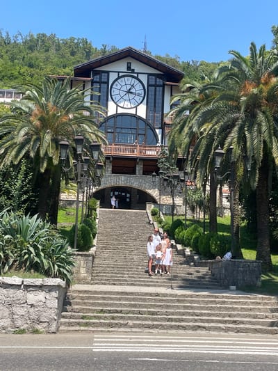 A family of five, two adults and three children, are posed on a wide, stone staircase leading up to a large, ornate building. The building, which features a prominent clock face with Roman numerals and ornate architectural details, is situated on a hillside covered in dense green foliage. Two tall palm trees flank the staircase, with one on the left and another on the right. A series of decorative lampposts are positioned along the side of the stairs. In the foreground, a portion of a paved road with a pedestrian crossing is visible. The sky is clear and blue, indicating a sunny day. The scene appears to be a tourist attraction in Gagra, Abkhazia.