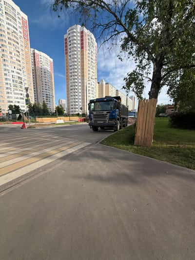 The image shows a street scene in Moskovskiy, Russia, on a sunny day. The foreground features a pedestrian crossing with yellow and white stripes, leading to an asphalt road. To the left of the road, there is a grassy area with a large tree providing shade. A blue SCANIA truck is parked on the side of the road. Behind the truck, a stack of wooden planks is leaning against a grassy mound.
In the background, several tall, modern residential buildings are visible. These buildings are characterized by their white facades with colorful accents of red and orange on the upper floors. The sky is blue with scattered white clouds.
The scene appears to be a typical urban environment, possibly a residential complex under construction or recently completed, given the presence of building materials. The time of day seems to be daytime, indicated by the bright sunlight and shadows. There are no people visible in the image.
Visible text on the truck includes "SCANIA" on the front and a license plate that appears to read "H856KX 793". A yellow triangular traffic sign with a black border and an image of a pedestrian is also visible on the left side of the road.