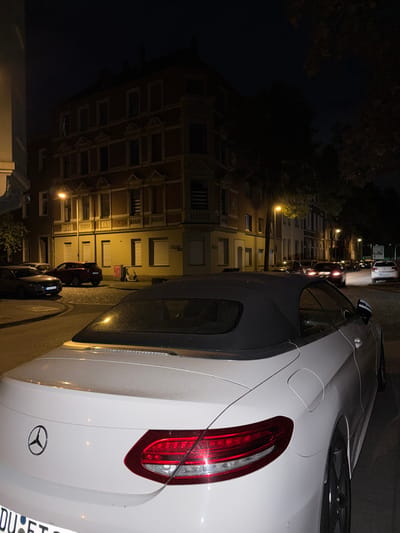 The image depicts a nighttime scene on a city street. A white convertible Mercedes-Benz is parked prominently in the foreground, its rear end facing the viewer. The car has its top down, revealing a black soft-top. The taillights of the Mercedes are illuminated. Behind the convertible, other cars are visible, some with their headlights on, creating streaks of light on the wet pavement. A red SUV is parked further down the street, next to a dark-colored sedan.
The background features multi-story buildings with distinctive architectural details, including ornate window frames and decorative elements. Streetlights cast a yellow glow on the buildings and the street. Trees with dark foliage are present, some of which appear to be bare or sparsely leaved, suggesting it might be autumn. An electric scooter is parked on the sidewalk near one of the buildings. A street sign is visible on a building in the middle ground, which, upon closer inspection, indicates "Bruckhausenstraße" and "Ruhrorter Straße," confirming the location as Duisburg, Germany, specifically the Bruckhausen area. The overall atmosphere is quiet and dimly lit, characteristic of a residential street at night. The pavement appears to be cobblestone in some areas.