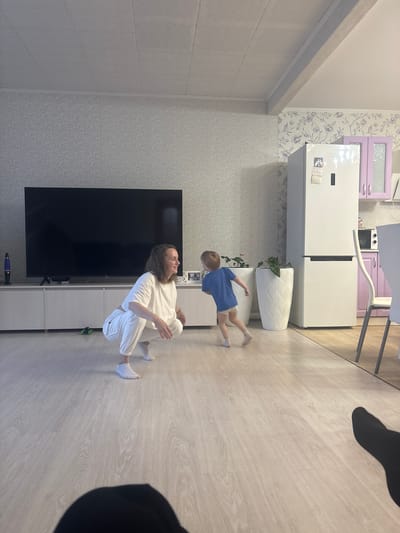 The image depicts the interior of a living space in Nestiukovo, Russia. A woman is sitting on the floor wearing a white top and white pants. A young child wearing a blue shirt and beige shorts is running near her. A large black television is mounted on the wall. A white refrigerator is visible in the background, along with kitchen cabinets in lavender and white. Two white planters are present next to the refrigerator. The floor appears to be light-colored wood laminate. There is patterned wallpaper on the walls. A vacuum cleaner is visible near the TV.