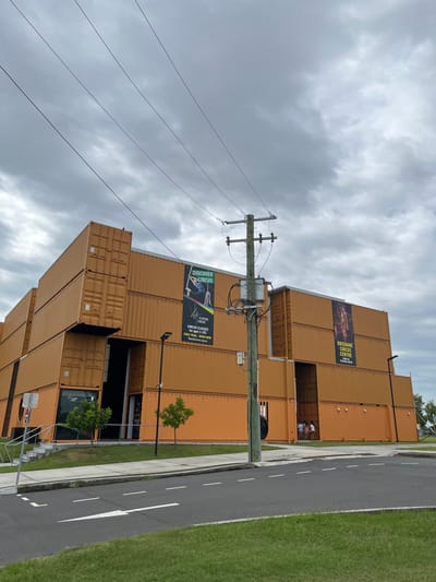 This image captures a multi-story building constructed from numerous stacked, warm orange-colored shipping containers, likely serving as a commercial or community center. The structure stands prominently under a largely overcast or cloudy sky.

On the right side of the building, a few individuals are visible through an open section on one of the upper levels, though their specific activity is not clear. In the foreground, a wooden utility pole with multiple cross arms and electrical wires runs vertically, with wires extending diagonally across the cloudy sky.

Adjacent to the building, a paved road with white dashed lane markings is visible, alongside a sidewalk and a strip of green grass. Two small trees with green foliage are planted along the sidewalk. Black street-style light poles are also present near the building.

Visible text on banners affixed to the container building provides context. A banner on an upper level reads "BRISBANE CIRCUS CENTRE." A second banner on a lower level features the text "DISCOVER CIRCUS" alongside an image of an aerialist, and details: "LYRA/TRAPEZE CLASSES for ages 5-18+ FREE TRIAL - BOOK NOW brisbanecircuscentre.com.au".

The scene depicts a daytime setting with cloudy weather, characteristic of an urban or suburban environment in Hamilton, Australia, consistent with the "Brisbane Circus Centre" identification.