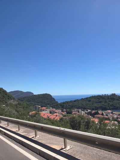 The image captures a scenic view of Novoselje, Montenegro, on a clear day. In the foreground, a metal guardrail runs horizontally across the bottom of the frame, separating a paved road from a vegetated slope. The road itself has a white line marking its edge. Beyond the guardrail, a cluster of buildings with red-tiled roofs are visible, nestled amongst lush green trees. The town appears to be situated on a hillside, with more greenery extending up the slopes. In the middle ground, rolling hills covered in dense forest lead towards the sea. The ocean is a deep blue, meeting the horizon under a vast, clear blue sky. A faint contrail is visible in the sky. The overall scene depicts a coastal town surrounded by natural beauty.