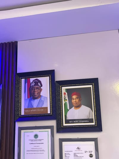 An indoor scene displays four framed items mounted on a light-colored wall, illuminated by overhead lighting that casts a blue hue on the upper left section and an adjacent vertical wooden slat panel.
The top row features two formal portraits in dark, ornate frames with gold accents. On the left is a portrait of Bola Ahmed Tinubu, identified as the President Commander-in-Chief of the Armed Forces of the Federal Republic of Nigeria, depicted smiling, wearing glasses, a light blue traditional attire, and a patterned cap. The background includes the Nigerian flag and coat of arms. To its right, a portrait of Senator Hope Uzodimma, identified as the Governor of Imo State, shows him looking forward, wearing glasses, a light-colored traditional top, and a red cap, with a similar Nigerian flag and coat of arms background.
The bottom row presents two framed business documents. The left document is a "Certificate of Incorporation" from the "Corporate Affairs Commission Federal Republic of Nigeria," dated July 5, 2023, for "J-AWAMSON BUSINESS SOLUTIONS LTD." The right document, partially visible, is a "Tax Clearance Certificate" from the Federal Inland Revenue Service (FIRS), also listing "J-AWAMSON BUSINESS SOLUTIONS LTD." The specific city location of Orie Uratta, Nigeria is not visually identifiable within the image; however, the displayed portraits of Nigerian political figures and corporate documents from Nigerian commissions firmly establish the national context.