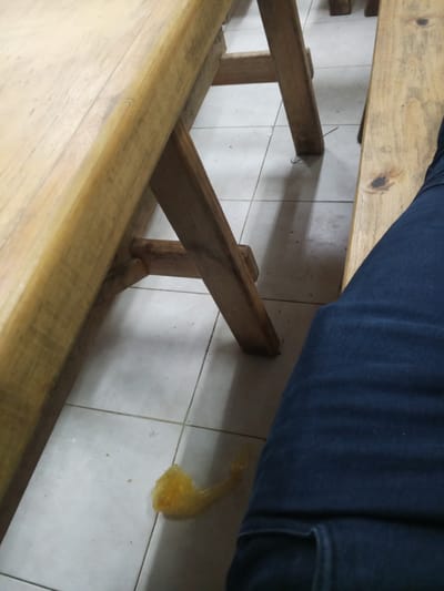 The image shows a close-up view of a wooden table and bench in what appears to be an outdoor setting in El Valle del Espíritu Santo, Venezuela. A person's dark blue jeans are visible on the right side of the frame, suggesting they are sitting on the bench. The ground is tiled, and a crumpled yellow plastic bag is lying on the floor near the leg of the table. The lighting suggests it is daytime.