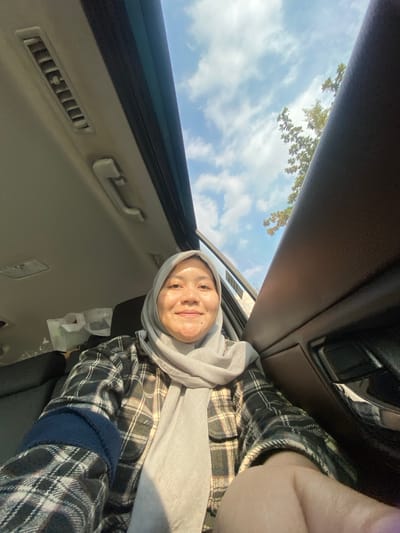 A young Indonesian woman wearing a light gray hijab and a black and white plaid shirt is taking a selfie inside a car. She is smiling and looking at the camera. The car appears to be in motion, as evidenced by the blurred trees and sky visible through the open sunroof and window. The interior of the car includes a black leather seat, a vent on the ceiling, and a door handle. The scene suggests the woman is traveling, possibly on a road trip or during her daily commute in Mandar Sari, Indonesia.