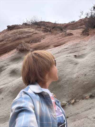 The image shows a person lying on a sandy, textured surface next to a rocky cliff. The person has blonde hair and is wearing a blue and white plaid shirt over a t-shirt with a nature-themed graphic. Sunglasses are visible on their head. The rocky cliff is reddish-brown and has some sparse vegetation growing from it. The sky appears overcast, suggesting a cloudy day. The scene is outdoors, with the sandy surface and rock formations indicating a natural, possibly coastal or desert environment. The person appears relaxed, looking upwards. There is no visible text in the image.