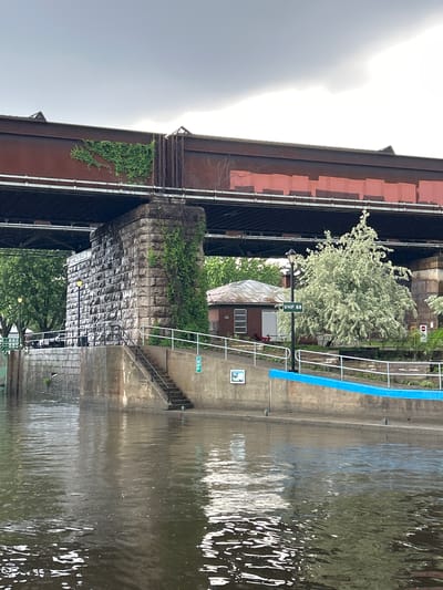 The image depicts a canal in Sainte-Anne-de-Bellevue, Canada, under a cloudy sky. A large railway bridge with rusted iron supports spans across the scene. The bridge's massive stone abutment on the right is partially covered by ivy. Below the bridge, a concrete walkway runs along the canal. A small brick building with a sloped roof and white-trimmed windows is situated on the walkway. A black lamppost with a green sign labeled "VHF 68" stands in front of the building. A light-colored, leafy tree grows near the building. On the left side of the canal, a stone wall rises from the water, with stairs leading up to the walkway. The canal water is murky and reflects the overcast sky. A blue barrier is visible along the edge of the walkway.
