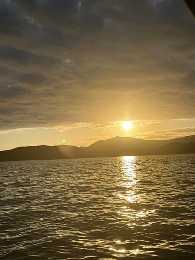 A vertical-format image, captured at sunrise or sunset, depicts a body of water in Dumbéa, New Caledonia, France, with a mountainous horizon. The sun, a bright golden sphere, is positioned slightly right of the frame's horizontal center, low above a distant, silhouetted landmass. Its intense light reflects across the water surface in a shimmering, vertical column extending towards the foreground. The sky transitions from a luminous golden hue at the horizon to darker, horizontally stratified grey clouds that occupy the upper half of the frame. A dark, undulating range of hills or mountains forms the horizon line, silhouetted against the bright sky. The water's surface exhibits minor ripples, enhancing the light reflection. No human figures, man-made structures, or identifiable objects beyond the natural environment are present.