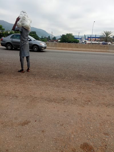 A person is standing on the side of a road in Kagini, Nigeria, carrying a large bag of what appears to be wheat flour on their head. The person is wearing a gray tunic and trousers, and open-toed sandals. A silver sedan is parked to their left on the paved road. The road is bordered by a concrete barrier. In the background, there are buildings, trees, and hills. The sky is overcast.