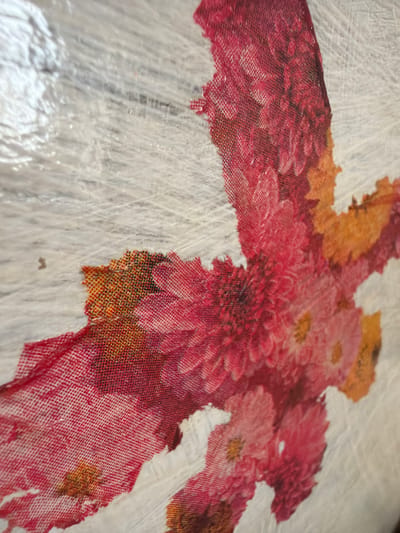 The image shows a close-up of a decorative element, likely a fabric or paper appliqué, featuring a floral design in shades of red, pink, and orange. The floral elements appear to be layered and have a textured, somewhat distressed appearance, with visible weave patterns in some areas. The background is a textured, off-white surface that resembles brushed paint or wood grain. The composition is abstract, focusing on the vibrant colors and textures of the floral motif. There are no people or identifiable landmarks present in the image, and therefore no specific location in Verkhnevilyuysk, Russia can be determined.