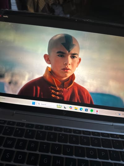 The media file presents an angled view of an open laptop, with the screen displaying content and a portion of the keyboard visible. The image itself is rotated counter-clockwise.

On the laptop screen, a young person is depicted, likely an actor from the live-action "Avatar: The Last Airbender" series, given the distinctive arrow tattoo on their bald forehead. They wear an orange and yellow robe, looking directly forward with a serious expression. The background of the displayed scene is light and somewhat indistinct, with hues of blue and white, suggesting an outdoor or expansive setting. The screen itself shows some light scuffs or dust.

Along the left edge of the screen, a taskbar or dock is visible, featuring several application icons. Prominently seen are the Netflix logo (with the word "Netflix" partially legible), Spotify, and other Microsoft application icons, including Edge. Towards the bottom right of the screen content, a search bar with a magnifying glass icon and the text "Q Search" is displayed. Below the Netflix logo, the fragmented text "ys ahead" can be seen.

To the right, a section of the laptop's dark gray keyboard is visible. Keys with numbers (2 through 9) and letters (such as Q, W, E, R, T, Y, U, I, O, P, S, D, F, G, H, J, K) are discernible, along with various symbols (!, @, #, $, %, ^, &, *, (, ), =, [, ]). Some function keys (fn) and an "insert" key are also apparent, with a subtle orange indicator light near the "insert" key. The setting appears to be indoors, focused on personal device usage.