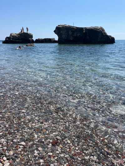 The image depicts a sunny day at a rocky beach in Zagrađe, Montenegro. The scene is split between a clear blue sky and the turquoise water of the sea, which laps against a shore covered in smooth, colorful pebbles. In the distance, large, dark rock formations rise out of the water, some of which are accessible by swimmers.
Several people are visible, enjoying the refreshing water and the opportunity for cliff diving. On one of the rock formations, three individuals are in the process of jumping into the sea, captured mid-air. Another person is in the water, swimming near the rocks. The clear water allows for a view of the pebbly seabed. The overall atmosphere is one of leisure and fun, characteristic of a summer day at the beach. The time of day is likely midday, given the bright sunlight and clear skies. There is no visible text in the image.