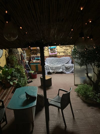 This image captures a nighttime view of an outdoor seating area, possibly a patio or garden, in Sabadell, Spain. The scene is illuminated by a string of small, orange lights draped across a thatched roof structure. In the foreground, a teal-colored table with decorative patterns sits between two dark chairs. On the table, a dark object, possibly a cap or a small bag, is placed. Behind this area, a couch covered with a patterned fabric is visible. To the right, a small olive tree with dense foliage stands next to a translucent partition. The ground is covered with reddish-brown tiles. In the background, shelves with various items, cleaning tools including brooms and buckets, and potted plants suggest a functional, lived-in space. The overall atmosphere is cozy and somewhat cluttered, hinting at a private residence or a small, informal gathering spot.
