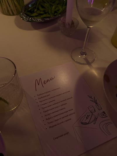 A dimly lit indoor scene features a white menu on a table. The menu has a handwritten-style title "Menu" in burgundy and a bulleted list of food items. Some of the visible items on the menu include "Хлебная корзина с сетом масел," "Крабовый риет," "Брускетта с печеным перцем и анчоусами," "Спаржa с хамоном," "Гранд салат с кейлом и печеной свеклой," "Трюфельный хумус с грибами," "Ассорти цветных томатов," "Вафли с креветочнм корни чизоm," "Тунец с пюре из цветной капусты," and "Морковь из печи с кремом." At the bottom of the menu, the text "Секретный десерт" is visible. To the right of the menu, a black line drawing depicts a table setting with a plate, napkin, knife, and small round food items. Further to the right, a sketch of a vase with branches and a glass is also present. The background shows a glass of white wine, a glass with ice and lime, and a dish of green edamame beans. The environment appears to be a restaurant setting, possibly in Chang Phueak, Thailand, given the provided context. There are no people visible in the image.