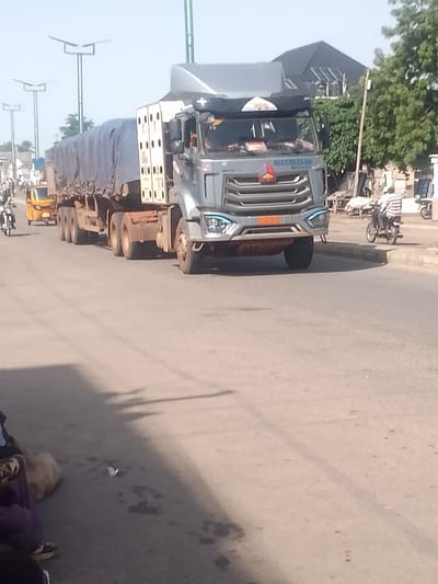 A grey articulated truck, its trailer covered with a dark blue tarp, drives on a paved road. The truck has a prominent grille with blue lights and a license plate that appears to read "AGBOM". A yellow auto-rickshaw is visible behind the truck, also on the road. Further in the background, there are buildings and trees. A person on a motorcycle, wearing a striped shirt, rides on the right side of the road, facing away from the camera. Several streetlights are visible on poles. The scene appears to be a daytime street view in a city.