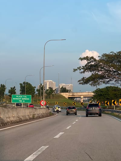 The image depicts a highway in Sepang, Malaysia, with multiple vehicles. A green sign indicates "PLAZA TOL BANDAR SERENIA 750 m". A speed limit sign displaying "5" is also visible. There are modern buildings in the background and a light blue sky with a visible contrail. Lighting poles line the side of the highway. Trees are present along the highway divider. The highway lane markings are white. Black and yellow chevrons mark the curve of the highway.
