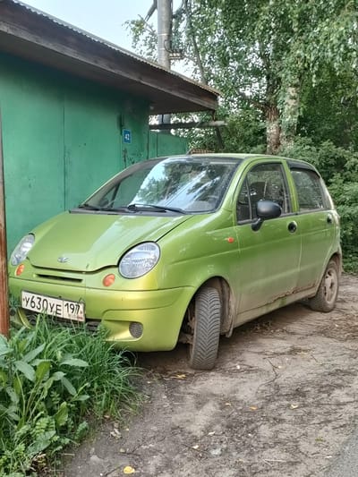 A green Daewoo Matiz is parked in front of a green garage in Boltino, Russia. The car is facing the viewer and is slightly angled to the left. The car is dirty and shows signs of rust on the lower part of the body. The license plate of the car is visible and reads "Y630YE 197". A number "42" is displayed on the garage door. Overgrown green grass is in front of the car, partially obscuring the lower bumper and license plate. The background features a large tree with green leaves and a wooden structure. The scene appears to be outdoors, with natural light indicating daytime. The weather seems to be overcast or late in the day, as the lighting is soft.