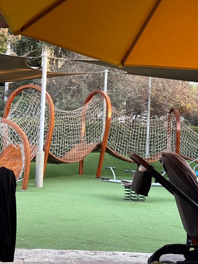 A view from underneath a yellow umbrella captures a playground or recreational area in San Isidro, Peru. Three wooden frame structures with white netting are visible, possibly components of a climbing structure or play apparatus, set on artificial grass. A stroller is present, partially obscuring the view and suggesting child activity. Beyond the playground equipment is an area of vegetation, including trees and bushes, which define the environment. The scene suggests a bright, outdoor setting suitable for children's activities. A portion of a person in black clothing appears at the bottom of the view.