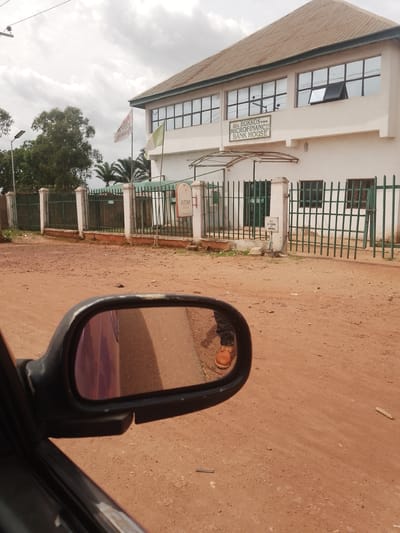 The image shows the exterior of a two-story building identified as the "BOKKOS MICROFINANCE BANK HOUSE" in Bokkos, Nigeria. The building is light-colored with a brown, corrugated metal roof. It features multiple large windows on both floors, some of which are tinted. A green metal fence encloses the property, with white-capped pillars along its length. An ATM machine is visible mounted on one of the fence pillars. Two flags are flying in front of the building; one is white with red lettering and the other is green. The ground in front of the bank appears to be unpaved and dusty, with some sparse vegetation and debris. In the foreground, a car's side mirror reflects the scene, showing a person's legs and feet wearing brown work boots and dark pants, indicating the photo was taken from inside a vehicle. The sky is overcast with clouds.