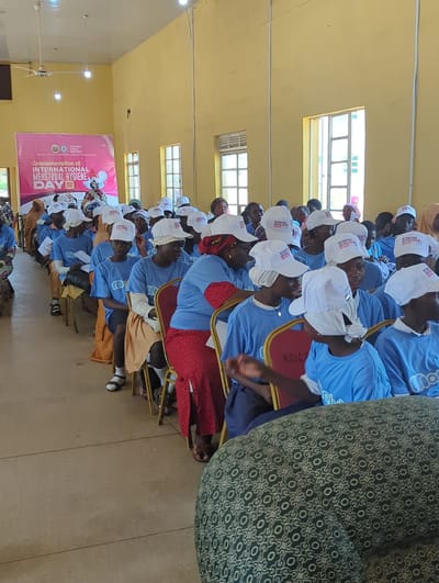 The image shows a gathering of people, likely at an indoor event. The attendees, primarily women and girls, are seated in rows, facing towards the front of the room. Many are wearing blue t-shirts and white caps with text that reads "INTERNATIONAL MENSTRUAL HYGIENE DAY". This indicates the event is a commemoration of this day.

A large pink banner with text in English is visible, further confirming the event's purpose. The banner reads, "Commemoration of INTERNATIONAL MENSTRUAL HYGIENE DAY". It also mentions "KADUNA STATE CO. MINSITRY OF EDUCATION" and "KADUNA PROJECT".

The setting appears to be a hall or meeting room with light-colored walls and large windows. A ceiling fan is also visible. The overall atmosphere seems attentive and engaged, with most individuals looking forward. The lighting suggests it is daytime. The presence of the banner and the specific theme point to an educational or awareness-raising event related to menstrual hygiene. The location context provided is Kakuri Gwari, Nigeria.