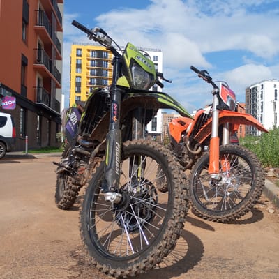 Two dirt bikes are parked on a dirt and paved surface in front of modern residential buildings. The building on the left is orange and white, with balconies. The building in the center is yellow and white, also with balconies. The building on the right is white. The dirt bikes are the main subjects, with the one in the foreground being black and green and the one behind it being orange. Both bikes are covered in mud, indicating they have been used off-road. The sky is blue with some white clouds. A white vehicle is parked to the left of the orange building, near a sign that says "OZON." The scene appears to be in an urban or suburban area.