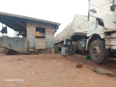 A worker is tending to a large, white truck with a covered trailer on a dirt road. Next to the road is a small, run-down building with a corrugated metal wall and a window displaying various items. A piece of wood debris lies in front of the building, and a green bucket sits near the truck. The scene is somewhat blurred, but the figures and objects are discernible.
