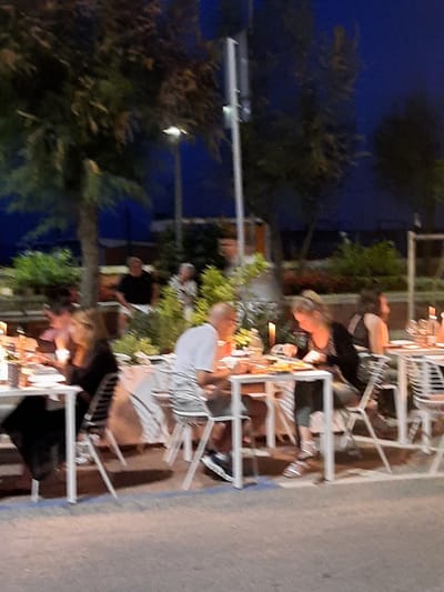 The image depicts an outdoor dining scene at dusk, likely in Pesaro, Italy, given the context. Several tables are set up, with people seated and interacting. One table in the foreground has a woman in a black top sitting, and another person to her left, whose face is not visible. Further back, a man in a white t-shirt is seated at a table, facing away from the camera. To his left, another woman with blonde hair in a black top is also seated at a table, looking down. Other individuals are present at various tables, some obscured by the low light and the outdoor setting. Trees and lighting fixtures are visible in the background, contributing to the evening ambiance. The ground appears to be paved. The overall impression is that of a casual outdoor restaurant or cafe.