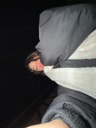 A person with brown hair is seen from a low angle, appearing upside down against a dark background. They are wearing a black jacket and a white, textured scarf or sweater underneath. The person is winking and has their lips pursed. A railing is visible slightly in the background. The lighting emphasizes the person's face, creating contrast with the surrounding darkness.