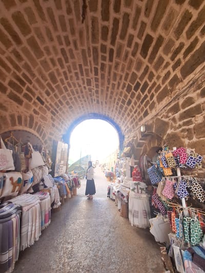 This is a forward-facing, eye-level, medium shot of a person walking through a covered market in Essaouira, Morocco. The market is enclosed by an arched brick ceiling and stone walls. The person is wearing a white long-sleeved shirt and a black skirt and is walking away from the viewer towards an opening of natural light. On either side of the walkway are market stalls displaying various textile goods. On the left, there are tote bags and pillows with embroidered designs. On the right, there are colorful pouches and small bags hanging from racks. The ground is paved with small stones and pebbles.