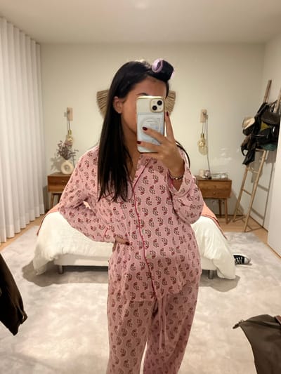 A single individual, identified as a young adult female, is depicted taking a mirror selfie indoors. The subject wears a two-piece pink pajama set featuring a repeating pattern of red and grey floral motifs and red piping along the placket. Her dark hair is styled with two purple hair rollers on the top of her head. She holds a gold-edged smartphone with a clear case in her right hand, obscuring her face, and her left hand rests on her hip. Her fingernails display dark red polish, and a ring is visible on her left ring finger.
The background shows a bedroom interior. A bed with a white duvet and a light-colored headboard occupies the central area. A light grey, textured area rug covers the floor. To the left of the bed, a wooden bedside table holds a vase containing dried flowers and a light-colored decorative object, with a wall-mounted lamp featuring a spherical bulb above it. To the right of the bed, another wooden bedside table is visible with a similar wall-mounted lamp. A large, circular, woven wall decoration is mounted above the bed. On the far right, a wooden ladder-style rack leans against the wall, supporting several bags and a hat. A pair of black low-top sneakers are present on the floor near the ladder. White pleated curtains are visible on the far left. The specific location within Lordelo do Ouro e Massarelos, Portugal, cannot be determined from the visual information provided in the image.