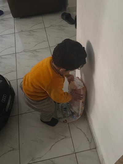 An indoor scene in Dar Bouazza, Morocco, depicts a young child from a high-angle perspective. The child, wearing a mustard-yellow long-sleeved top and light grey sweatpants, is in a crouched position, facing towards a white interior wall. Their dark, curly hair is visible. Both hands are placed on a large, clear plastic-wrapped package of disposable diapers, which is resting against the wall. The diaper package displays the number "38" and other partially legible branding. The floor is composed of light-colored square tiles with dark, irregular veining patterns. In the upper left background, a black object with a silver "AUDI" logo is visible, alongside a dark slipper and a piece of brown furniture. Shadows are cast on the white wall to the child's right.