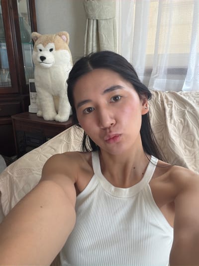 A person is lying on a bed covered with a beige patterned comforter. The person is wearing a white ribbed tank top. Their dark hair is loose around their face and shoulders. To the left of the person, on a dark wooden surface, is a stuffed animal resembling a husky. In the background, there is a window covered with translucent blinds and light-colored curtains. The image's location is likely within an indoor residential setting; there is no way to discern the Kawasaki, Japan location based on the image details.