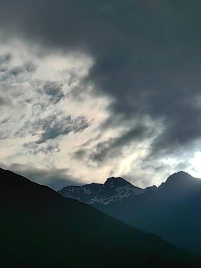 The image depicts a dramatic mountain landscape under a heavily clouded sky. In the foreground, a dark, silhouetted mountain slopes upward from the bottom left, obscuring much of the lower portion of the scene. Further back, a range of jagged mountain peaks emerges, some capped with snow, against the turbulent sky. The clouds are a mix of dark, ominous grays and lighter, illuminated patches, suggesting either an approaching storm or the aftermath of one. A bright, diffused light emanates from behind the central peaks, casting a soft glow through the clouds. The overall impression is one of rugged, untamed natural beauty. The location is specified as Kardang, India. No people or specific objects are visible in the image, only the natural elements of mountains and sky.