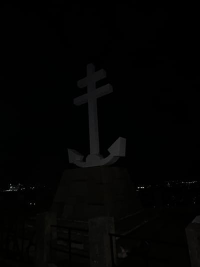 The photograph shows a stylized anchor sculpture mounted on a dark structure, possibly a pier or dock, in an outdoor setting. The anchor is light-colored, contrasting with the surrounding darkness. It is placed off-center, positioned towards the left side of the frame. The structure onto which the anchor is mounted appears to be made of concrete or stone, and it is significantly darker. Parts of a building are visible behind. There are water droplets falling in the frame near the top right corner, suggesting a damp environment. The setting appears to be Gourock, United Kingdom.