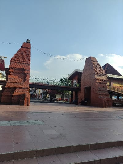 The image depicts a red brick structure, possibly a remnant of a historic building, located in Mojokerto, Indonesia, at coordinates -7.464 latitude and 112.432 longitude. The structure is composed of two tall, rectangular brick pillars, each with a distinct, slightly angled shape. A metal bridge, with a black railing, connects the two pillars, extending over a paved area. The bridge appears to be part of a pedestrian overpass. The structure is in a state of partial disrepair, with some of the bricks missing or cracked. There are several individuals, dressed in casual attire, standing or walking on the paved area beneath the bridge. The sky is a light blue, with white clouds scattered across it. The ground is composed of a mixture of grey concrete and red brick pavers. Green foliage is visible in the background. The scene is illuminated by natural daylight, suggesting a daytime setting.