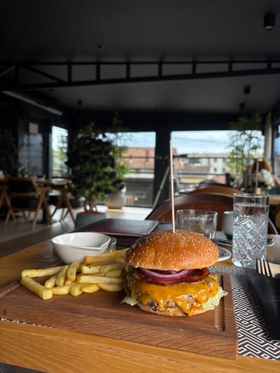 A close-up, eye-level shot captures a gourmet burger and fries served on a wooden board at a restaurant. The burger, topped with sesame seeds, features a thick patty, melted cheddar cheese, red onion rings, and a slice of bacon. A skewer sticks out of the top of the bun. A generous portion of golden french fries is arranged to the left of the burger, alongside a small white dish, likely for dipping sauce. In the background, out of focus, are two glasses of water, one with a decorative pattern, and a black napkin with the word "Presido" visible. The setting appears to be a contemporary restaurant with large windows offering a view of an urban landscape, possibly Loznica, Serbia. The interior features dark ceilings with exposed structural elements and what appear to be artificial plants. The overall lighting suggests daytime, with natural light filtering through the windows. There are no discernible people in the immediate foreground, but hints of patrons and outdoor scenery are visible in the background.