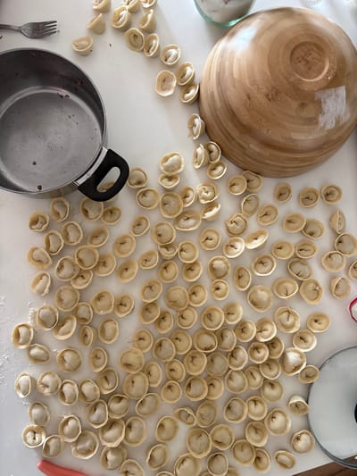 A top-down, medium shot captures a white countertop laden with uncooked pelmeni. The dumplings, uniformly shaped with pleated edges, are scattered across the surface in a dense arrangement. To the left, a large, unfinished wooden bowl is partially visible, its circular grain texture evident. In the lower left quadrant, a stainless steel pot with a black handle sits prominently. A metal fork rests near the pot, and a few small, dark crumbs are scattered on the countertop.

The scene appears to be indoors, on a well-lit kitchen counter, suggesting a domestic setting. The natural light implies daytime. There are no people visible, but the presence of food preparation items indicates an activity related to cooking or meal assembly. The pelmeni are the primary focus, showcasing their delicate, handmade appearance. The overall impression is one of a culinary endeavor, possibly the preparation of a traditional dish. No text is visible in the image.