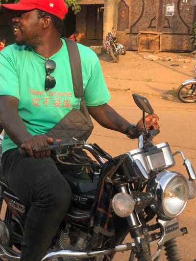 An individual, identified as male with dark skin, is seated on a motorcycle on an unpaved, reddish-brown dirt road in Neni, Nigeria. He is shown from the waist up, wearing a red baseball cap with "UR" on the front, a bright green short-sleeved t-shirt displaying "Never Compromises" in red English text and accompanying Chinese characters (从不妥协). Black sunglasses are hung from his t-shirt collar. He has a black, quilted shoulder bag slung across his chest and a dark wristwatch on his right wrist. His hands are positioned on the motorcycle's handlebars, with the right hand near the throttle/brake lever. The motorcycle is a black Kymstone CG125 model, featuring chrome handlebars, a prominent chrome headlight, a smaller round amber indicator light on the left, and a black side mirror on the right. A blue, white, and orange/red checkered cloth is draped over the handlebars.
In the background, a large dark metal gate with decorative elements is visible, serving as an entrance to a structure. A motorcycle is parked upright near the gate. An overturned wooden table or bench lies on the ground to the right of the gate, with a light-colored, indistinct object nearby. The front wheel of another motorcycle is partially visible on the far right. The scene is brightly illuminated by natural sunlight, indicating daytime conditions.